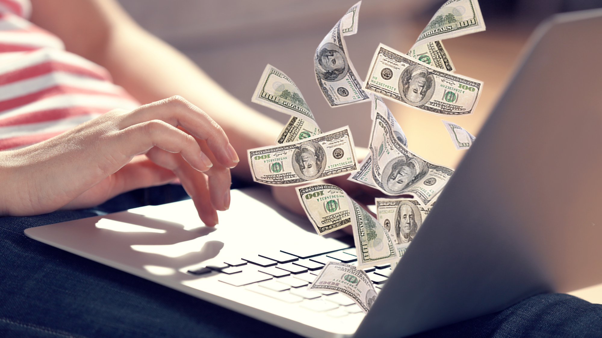 A laptop on a lap with 100 dollars flying