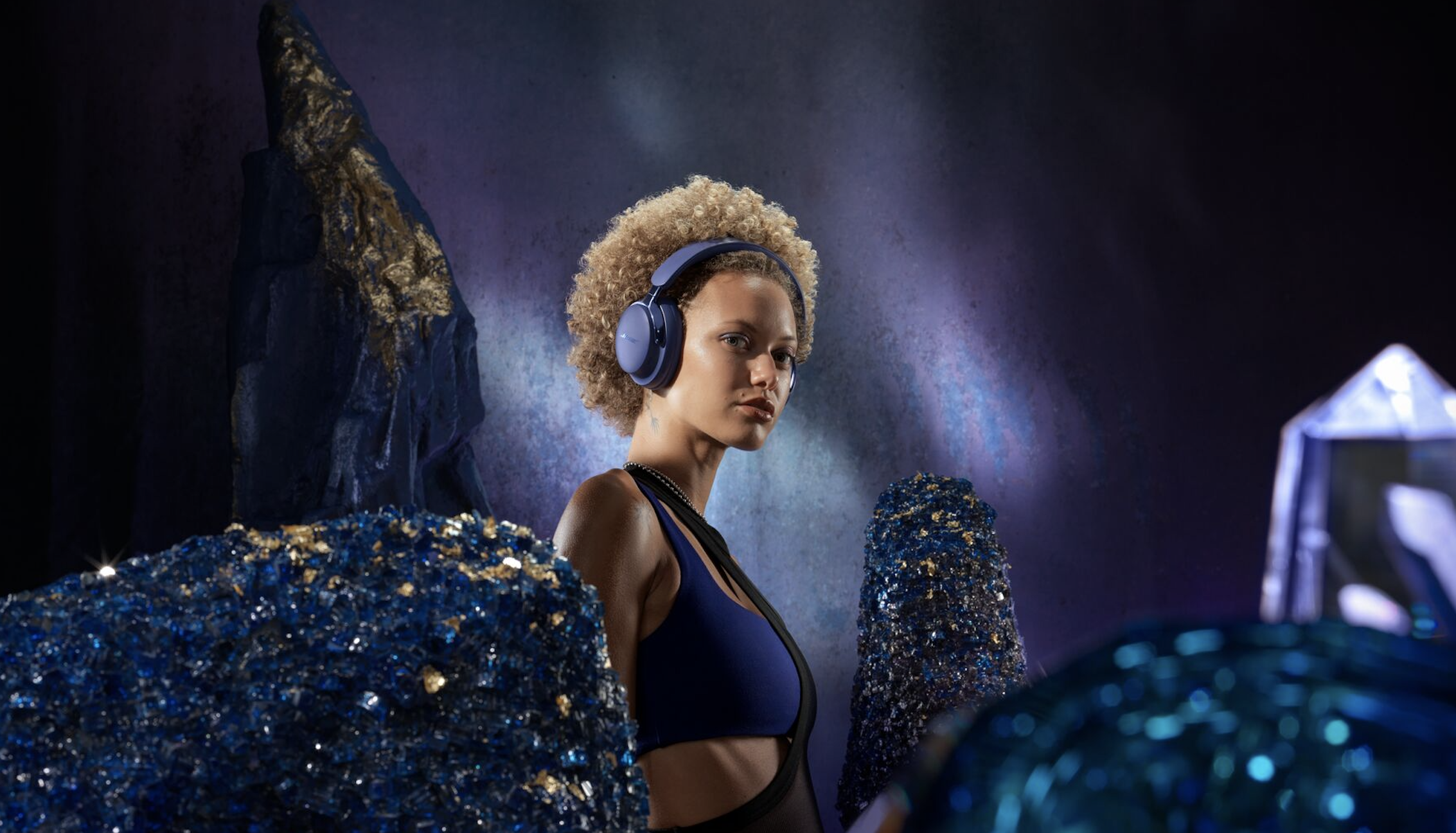 Bose qcu (2nd gen) headphones in 'Midnight violet' used by a woman, surrounded by purple crystals