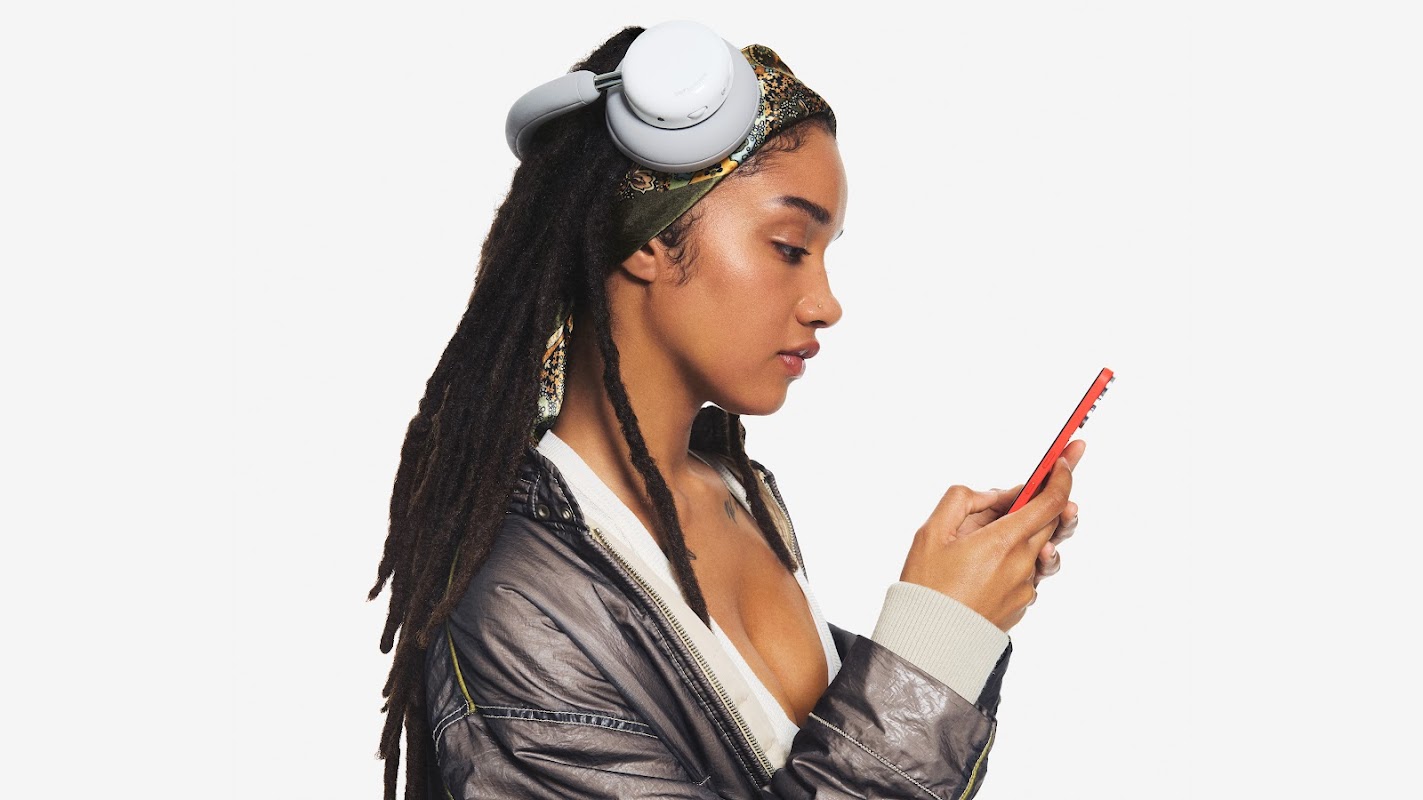 A young woman with black braided hair with a white lid and a silver jacket looks at a red phone. She has a handkerchief on her head and a pair of CMF headphones Pro are parked on him.