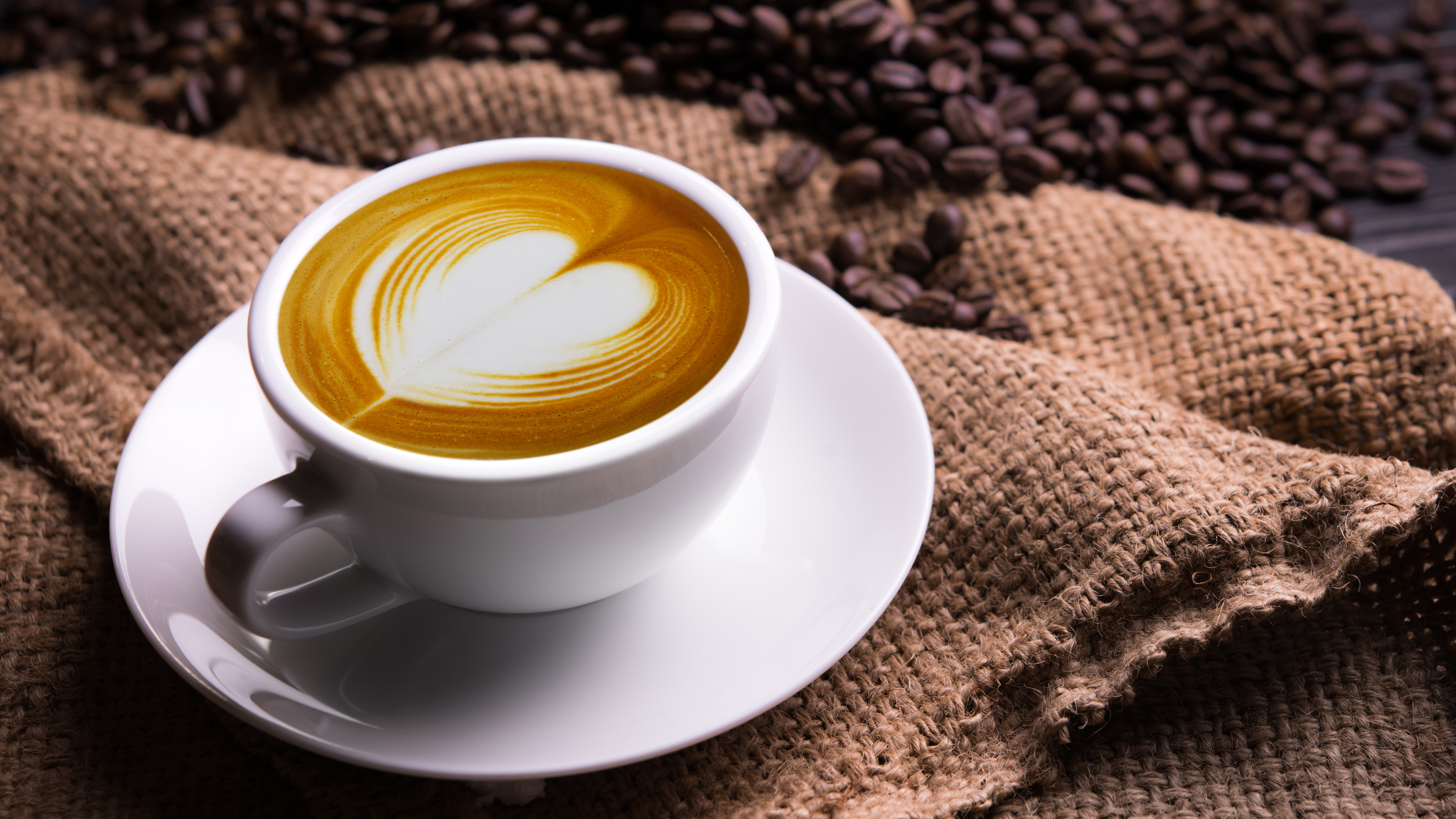 A coffee with milk art at the top in a cup of white coffee and saucer next to a hessian sack and coffee beans