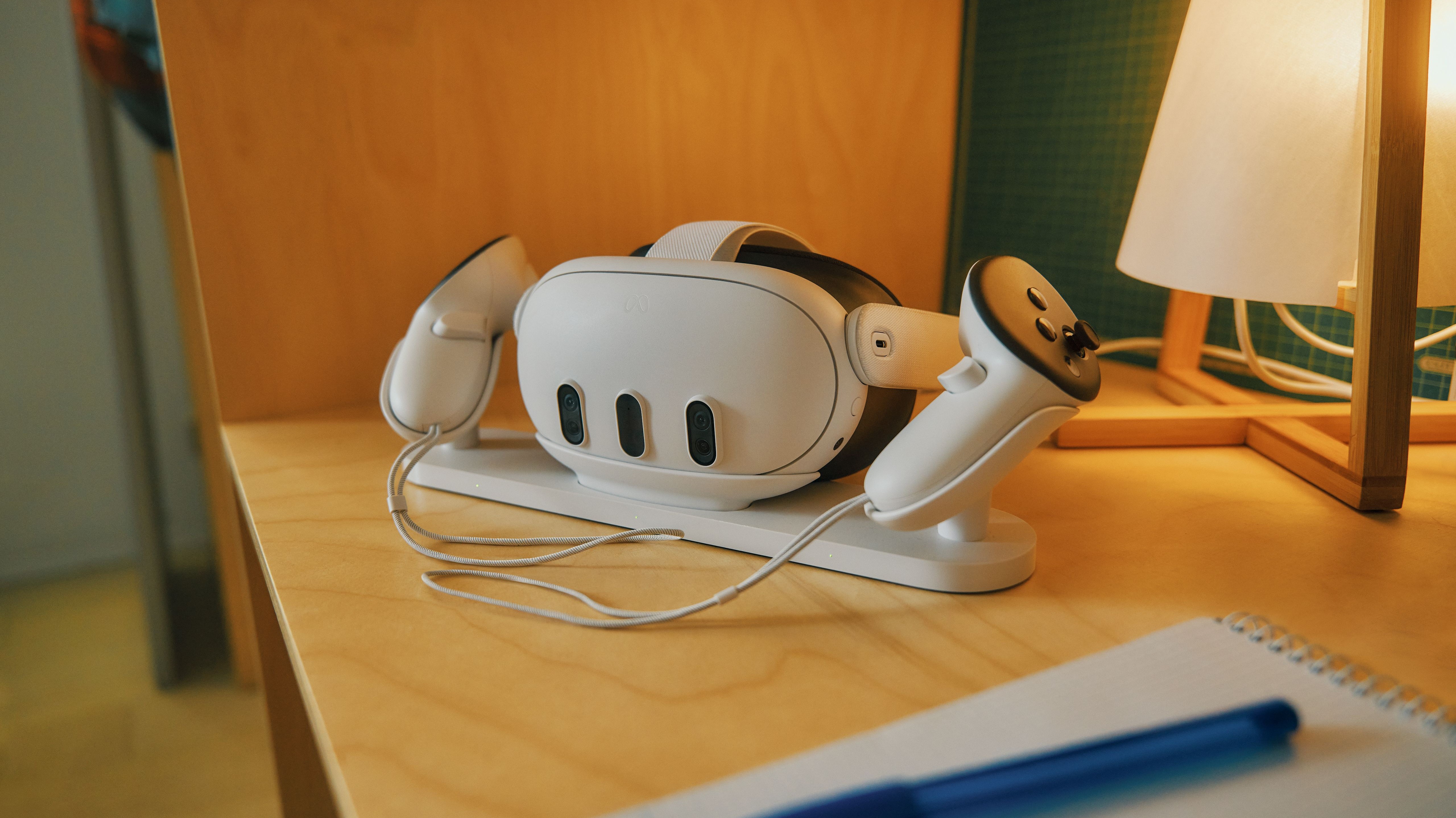 The Meta Quest 3 and controllers in their charging station, which sits on a wooden desk next to a lamp.