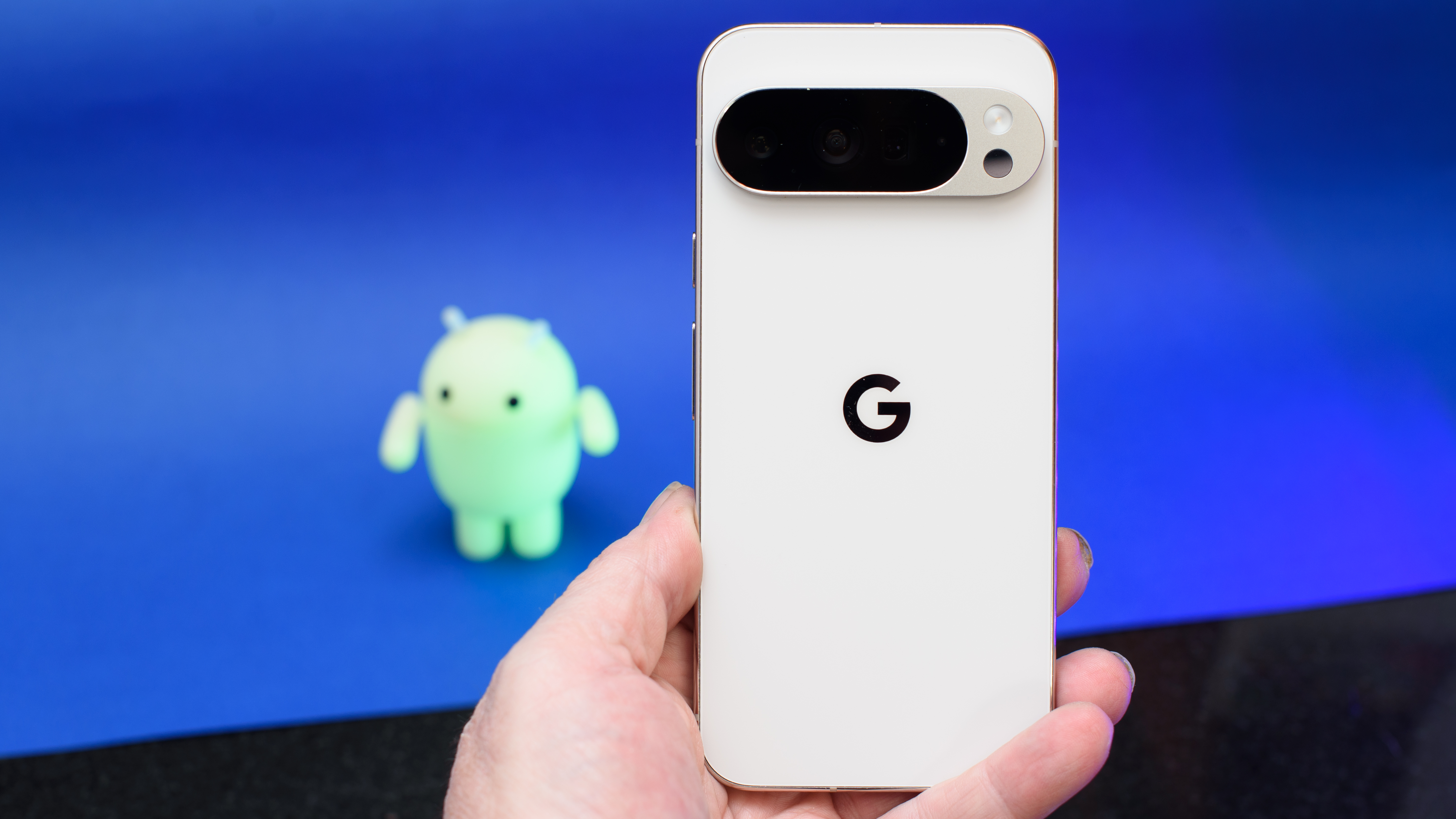 Google Pixel 10 Pro XL in porcelain on a blue background with a green neon Android figure in the background