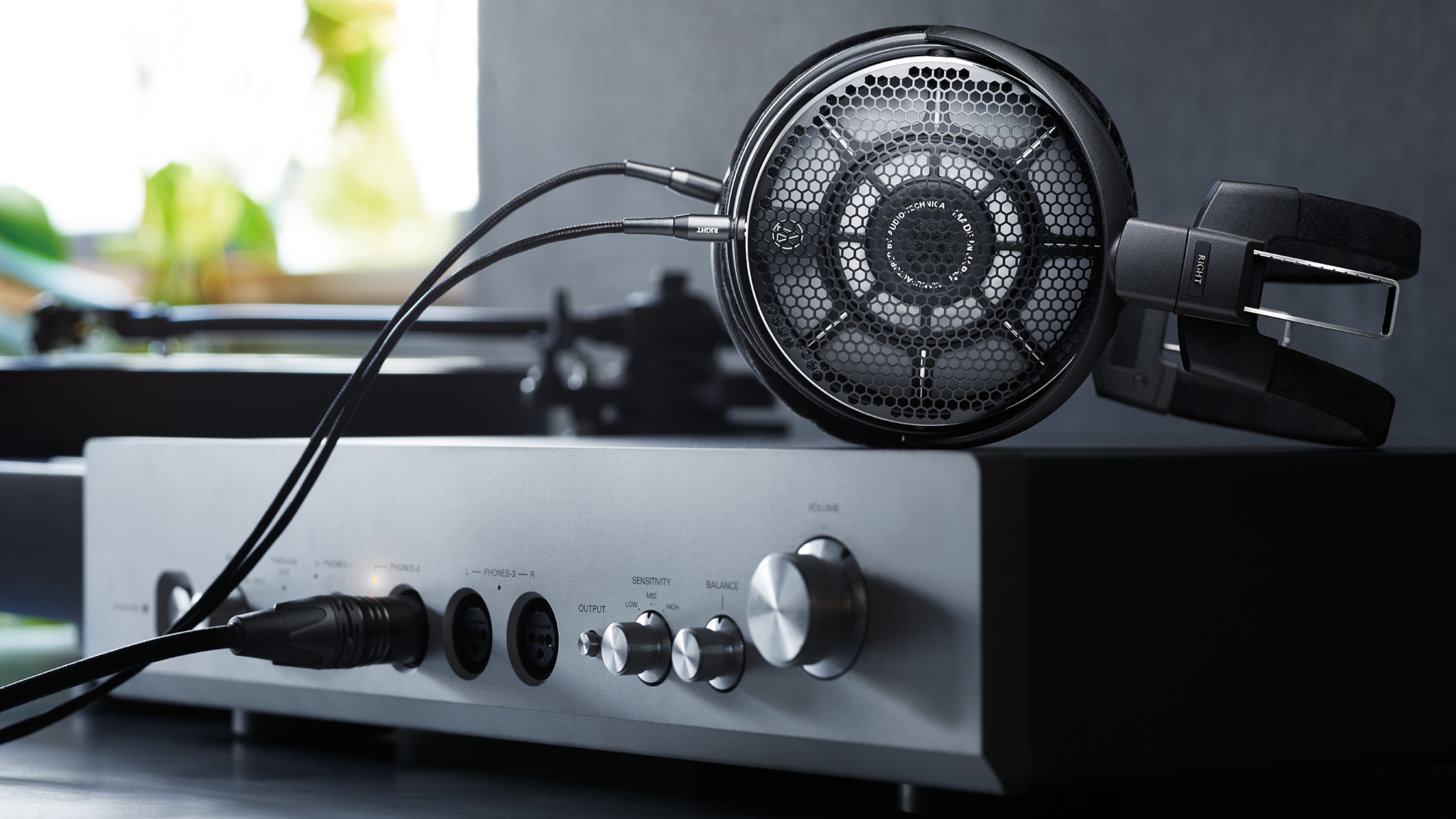 Product photo of Audio-Technica ATH-ADX7000 over-ear headphones on top of a headphone amplifier with a window in the background