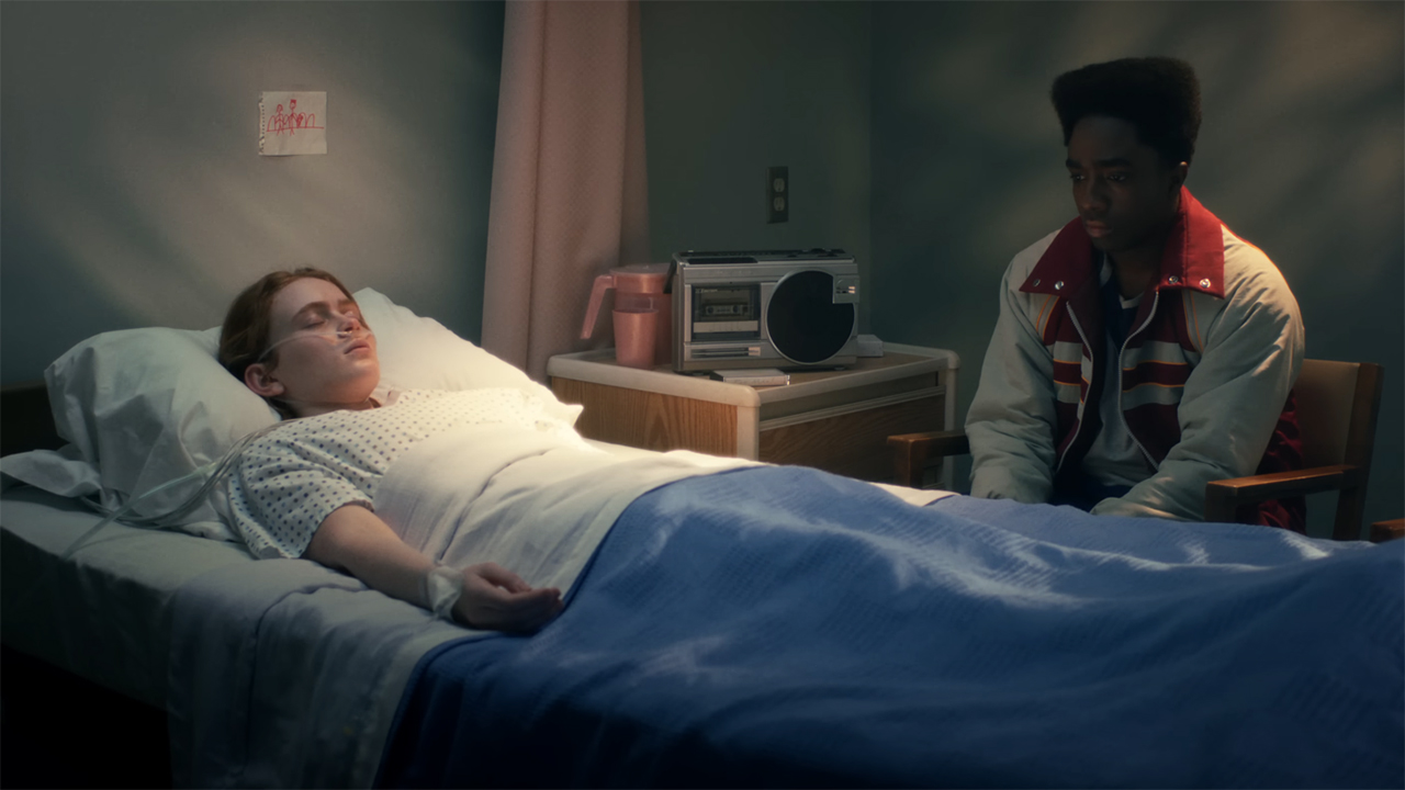 A comatose Max lying in a hospital bed while Lucas sits next to him in season 5 of Stranger Things.