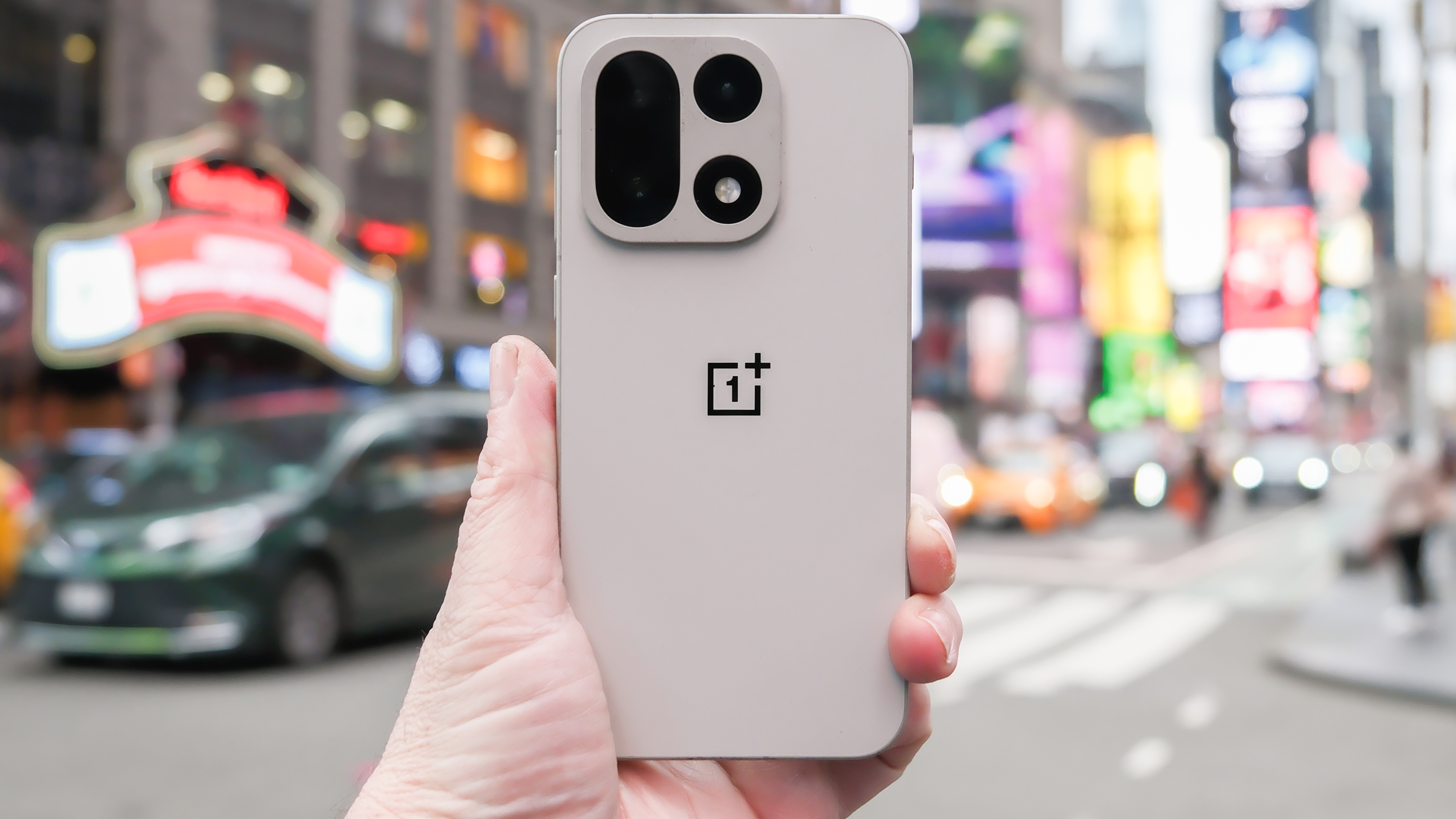 OnePlus 15 in Sand Storm color held in hand with cameras and back shown in the middle of a Times Square intersection in New York City in front of the Hard Rock Hotel. The background is blurred with cars and people passing by.