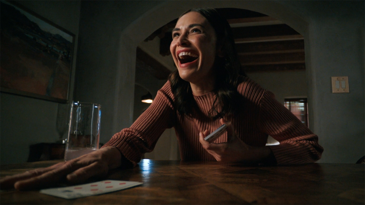 Zosia smiles widely and places her right hand on a stack of board cards in episode 8 of Pluribus.