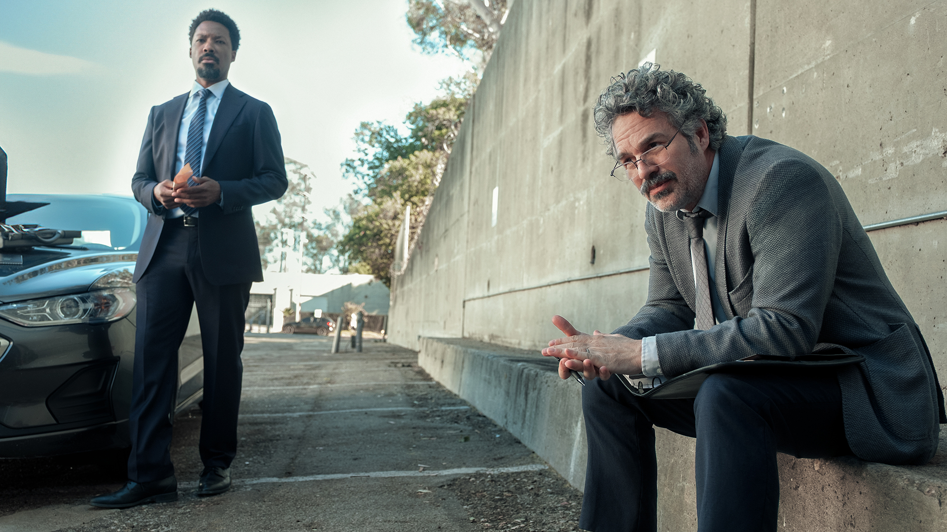 Detective Tillman stands next to a car while Detective Lubesnick sits on a nearby wall in Crime 101.