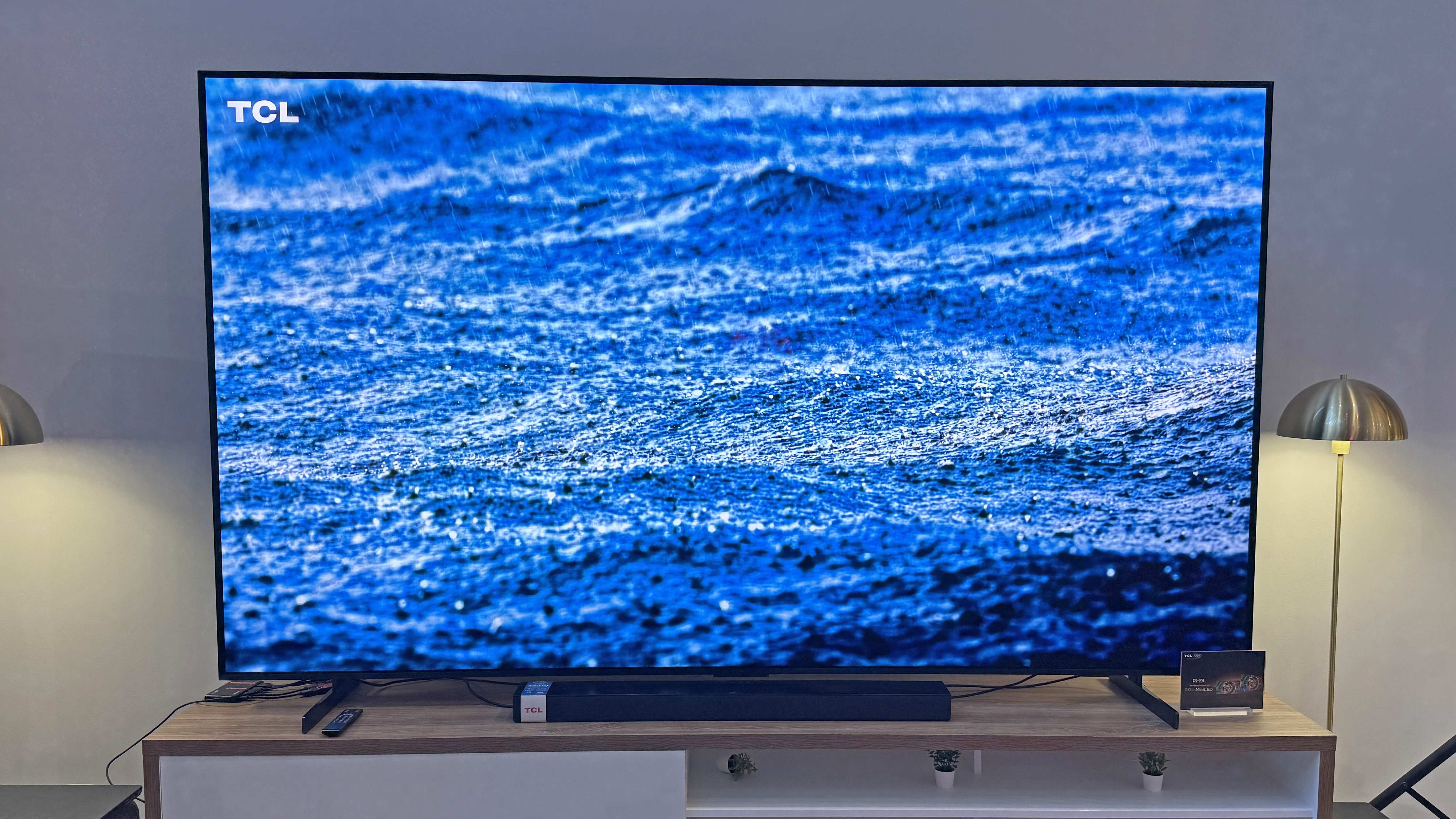 The TCL RM9L TV at a launch event, showing an image of rain falling over the ocean, with a striking blue tone and lots of detail in the rain splashes.