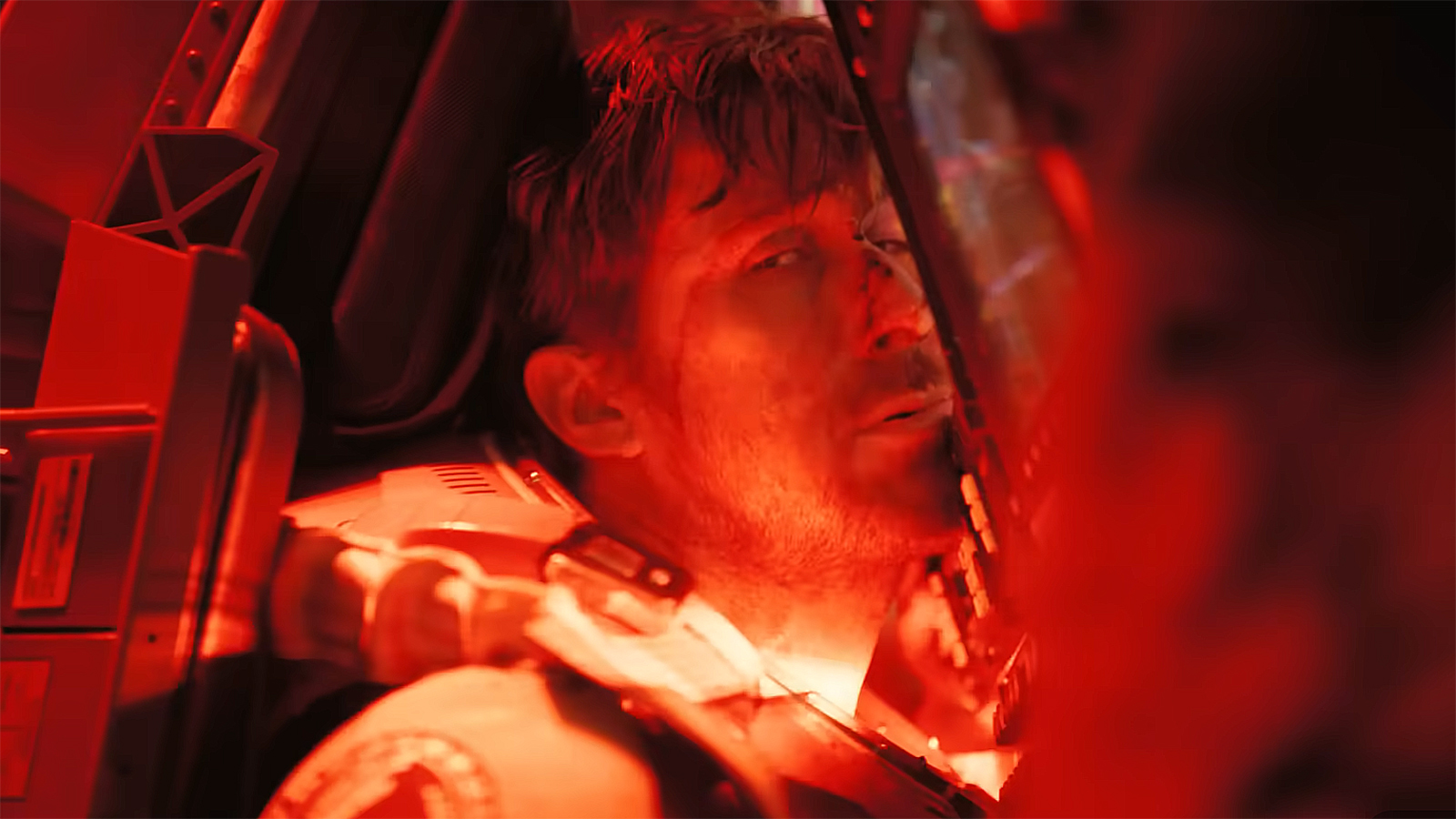 An injured Ryland Grace sits in a seat pressed against the console of a red-lit spaceship in Project Hail Mary.
