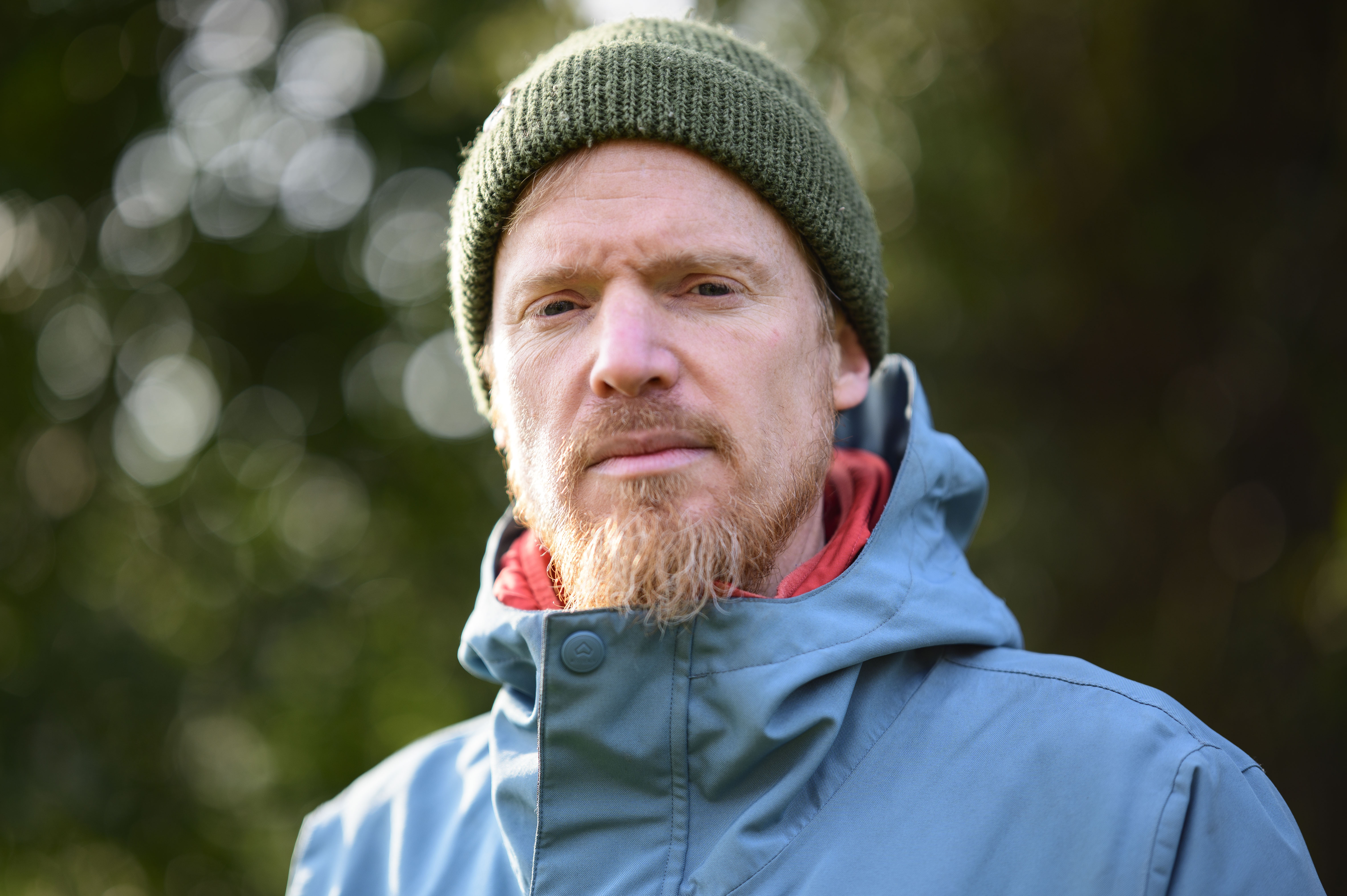 Portrait of a man in a blue coat and a green hat, with dappled light through a tree in the background