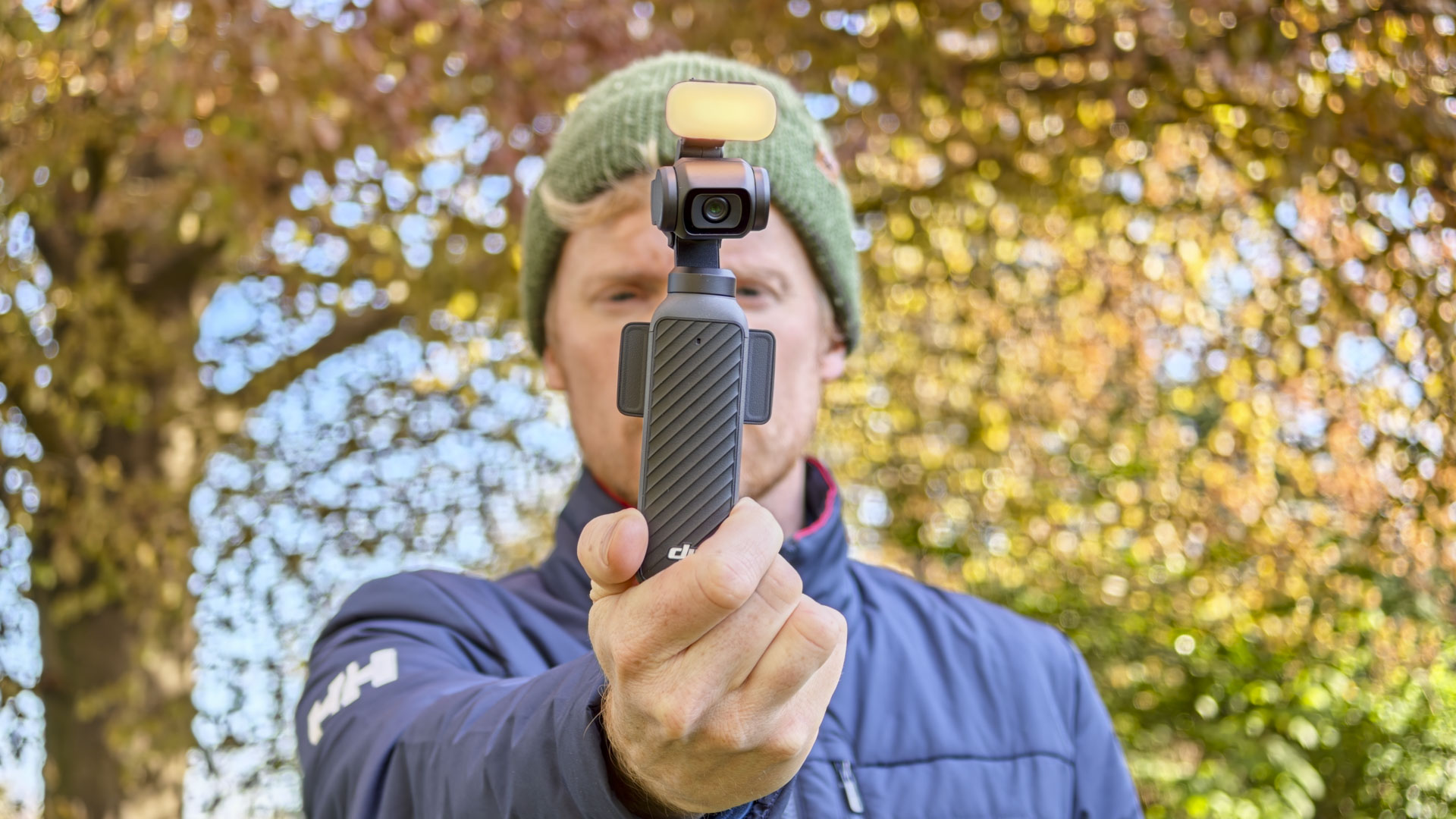 Man in beanie holding DJI Osmo Pocket 4 vlogging camera with its magnetic fill light, an autumn tree behind him