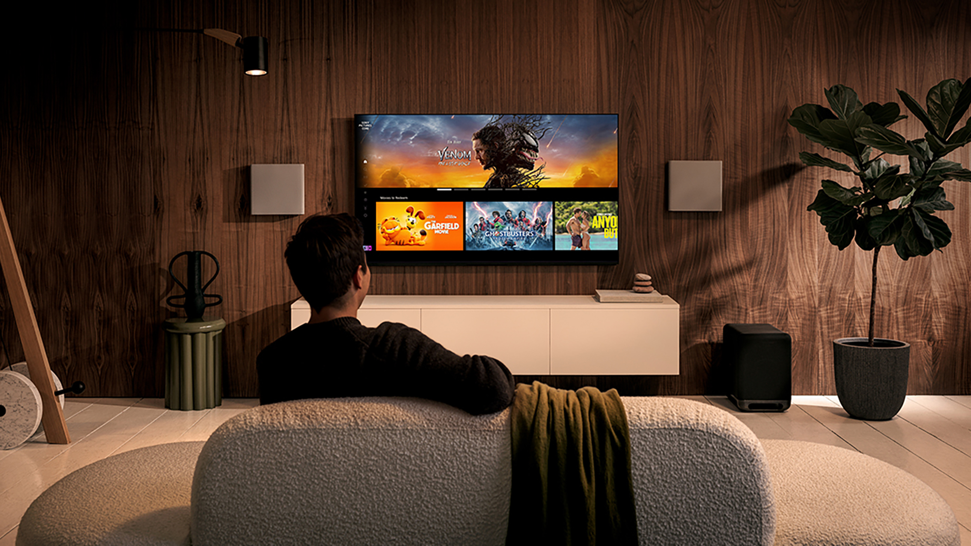 A man seen from behind, sitting on a sofa watching a television, which shows thumbnails of the selected content.