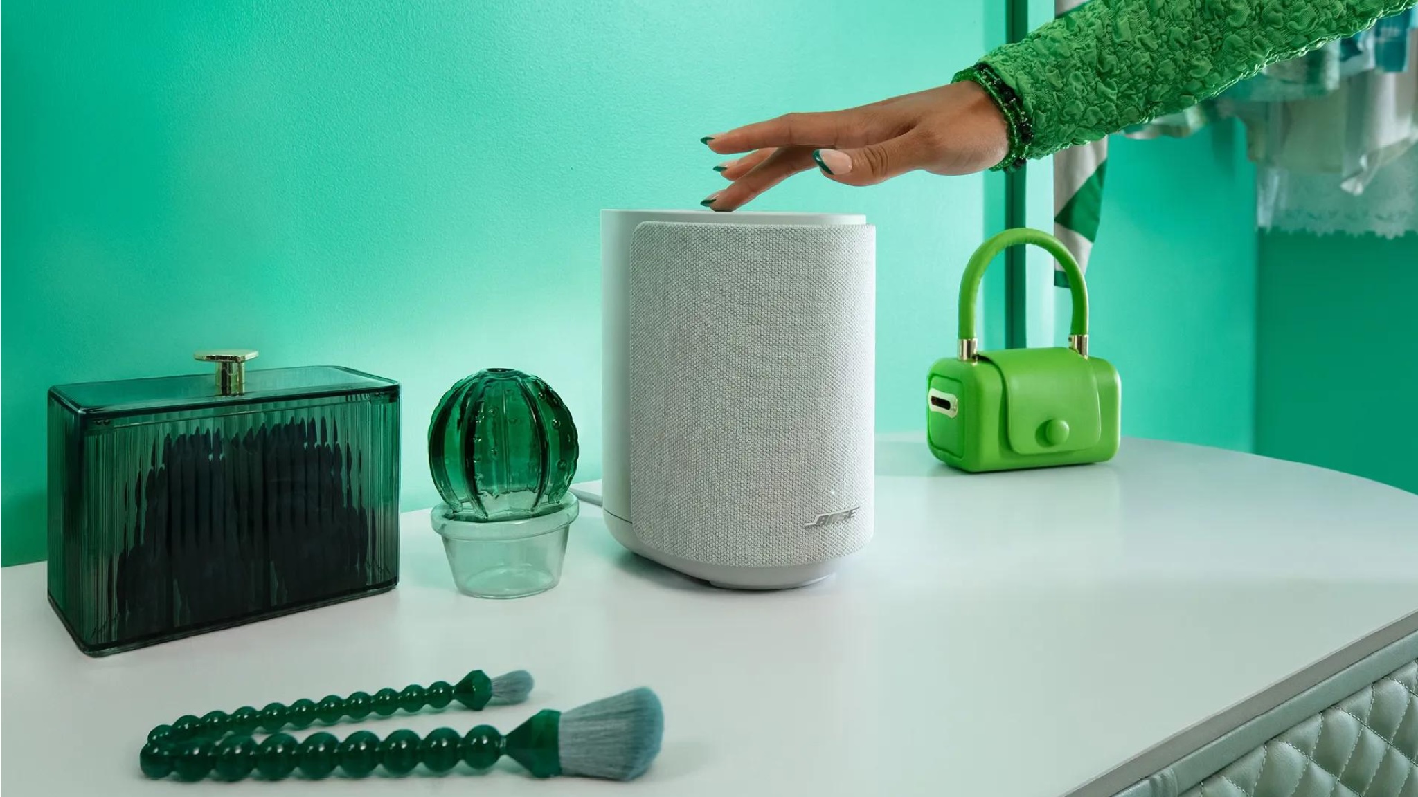 Bose Lifestyle Ultra speaker on a white surface in front of a green wall. There are green decorations on the white surface and a person's hand touches the top of the speaker.