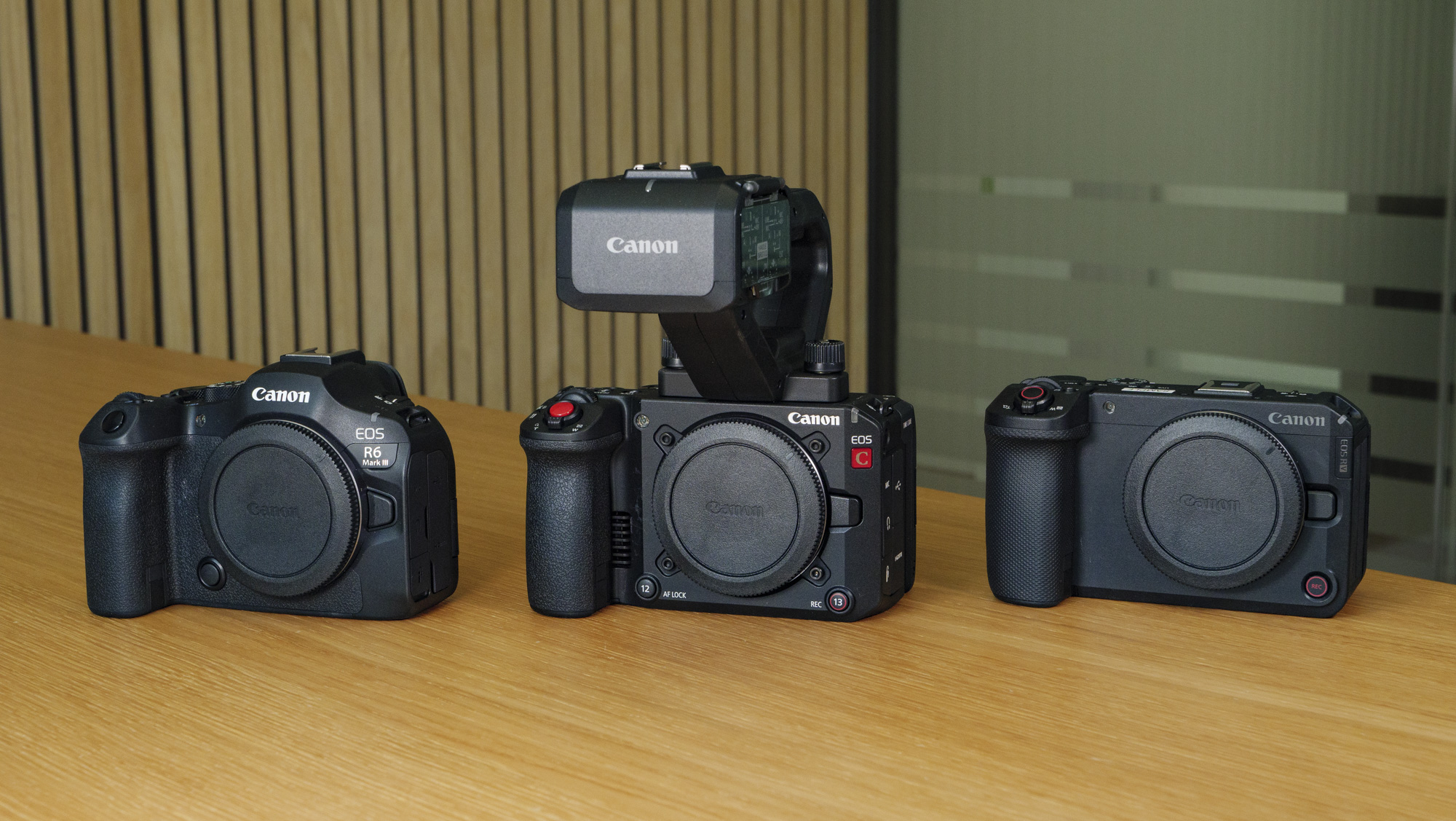 The Canon EOS R6 V on a wooden table next to the Canon EOS R6 III and the EOS C50