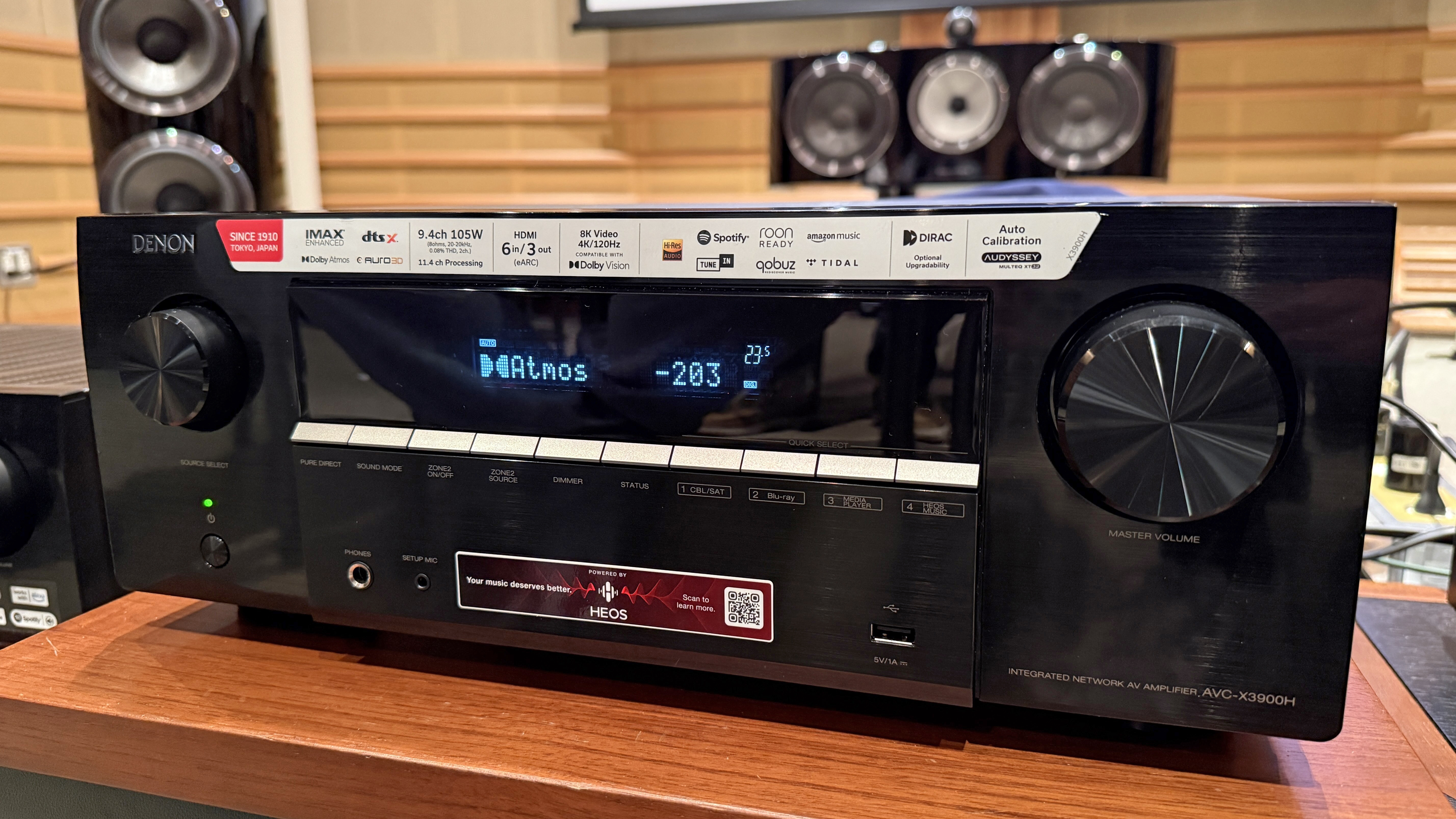The Denon AVR-X3900H with Bowers & The Wilkins speaker is barely visible behind him. It's a black box with some dials and buttons on the front. The screen says it is playing a Dolby Atmos signal.