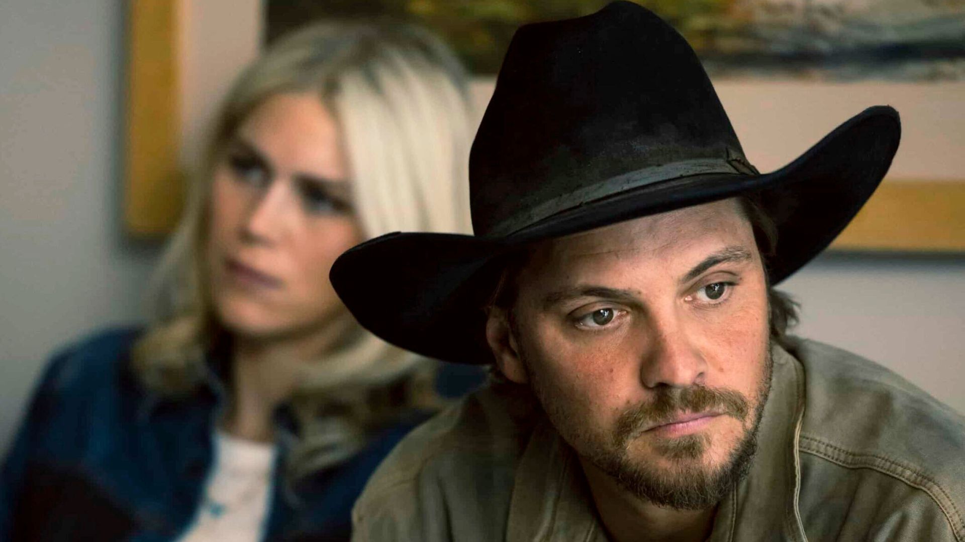Kayce looks into the distance as a blonde woman sits behind him.