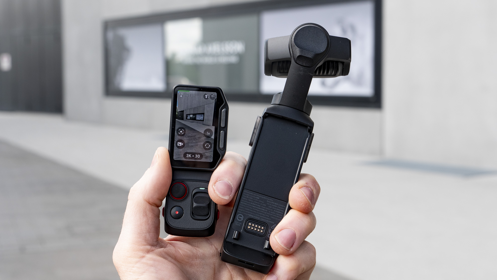 Hand holding Insta360 Luna Ultra vlogging camera and its screen, outdoors in a gray urban scene