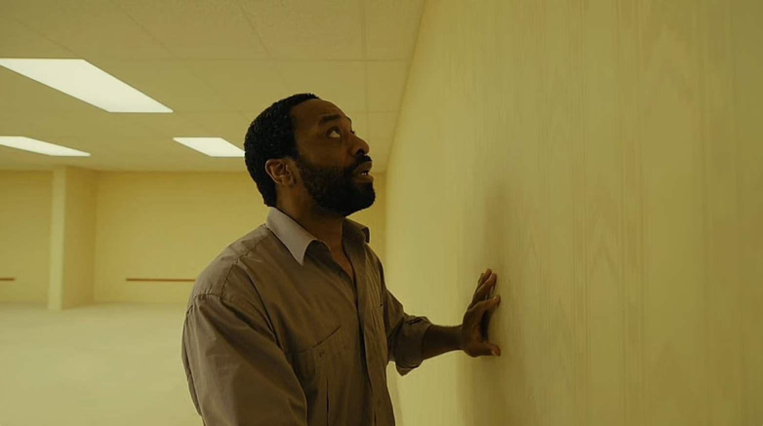Chiwetel Ejiofor in Backrooms, where he is in an empty yellow room. He is touching a wall and looks scared.