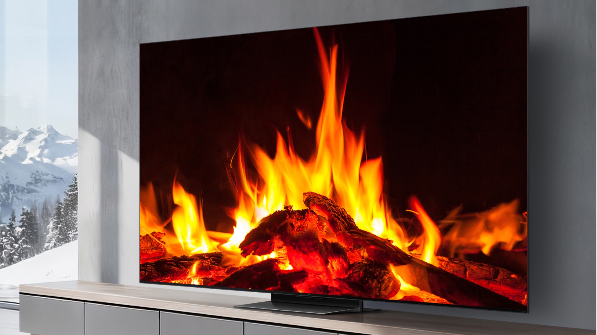 A TCL C7L television showing a wood fire. You are in a gray decorated room with snow-capped mountains visible through a full-height window to the left of the television unit.