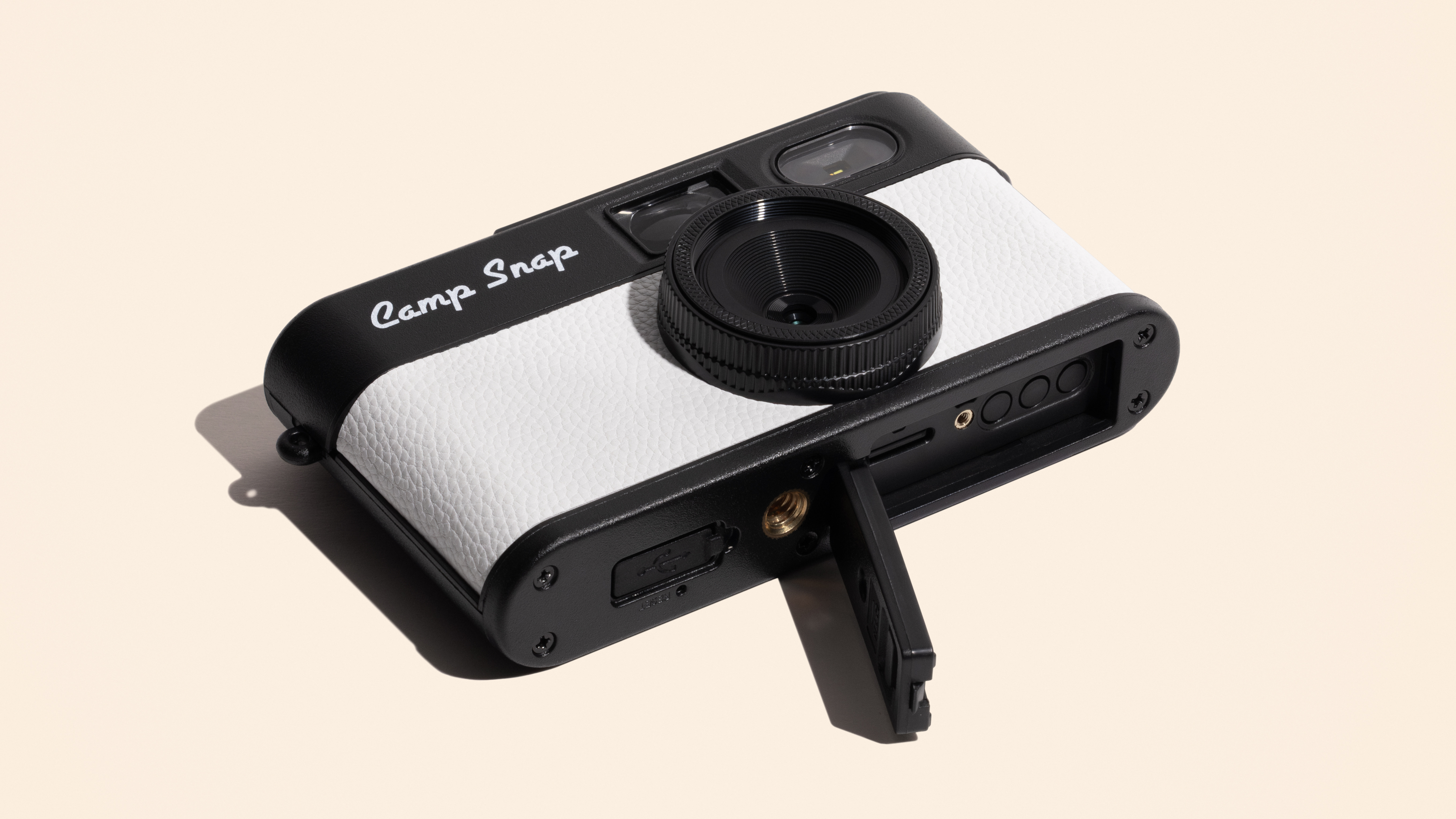 Photograph of the small Camp Snap 2 compact digital camera in white, with the bottom plate showing the open memory card door and threaded tripod mount.