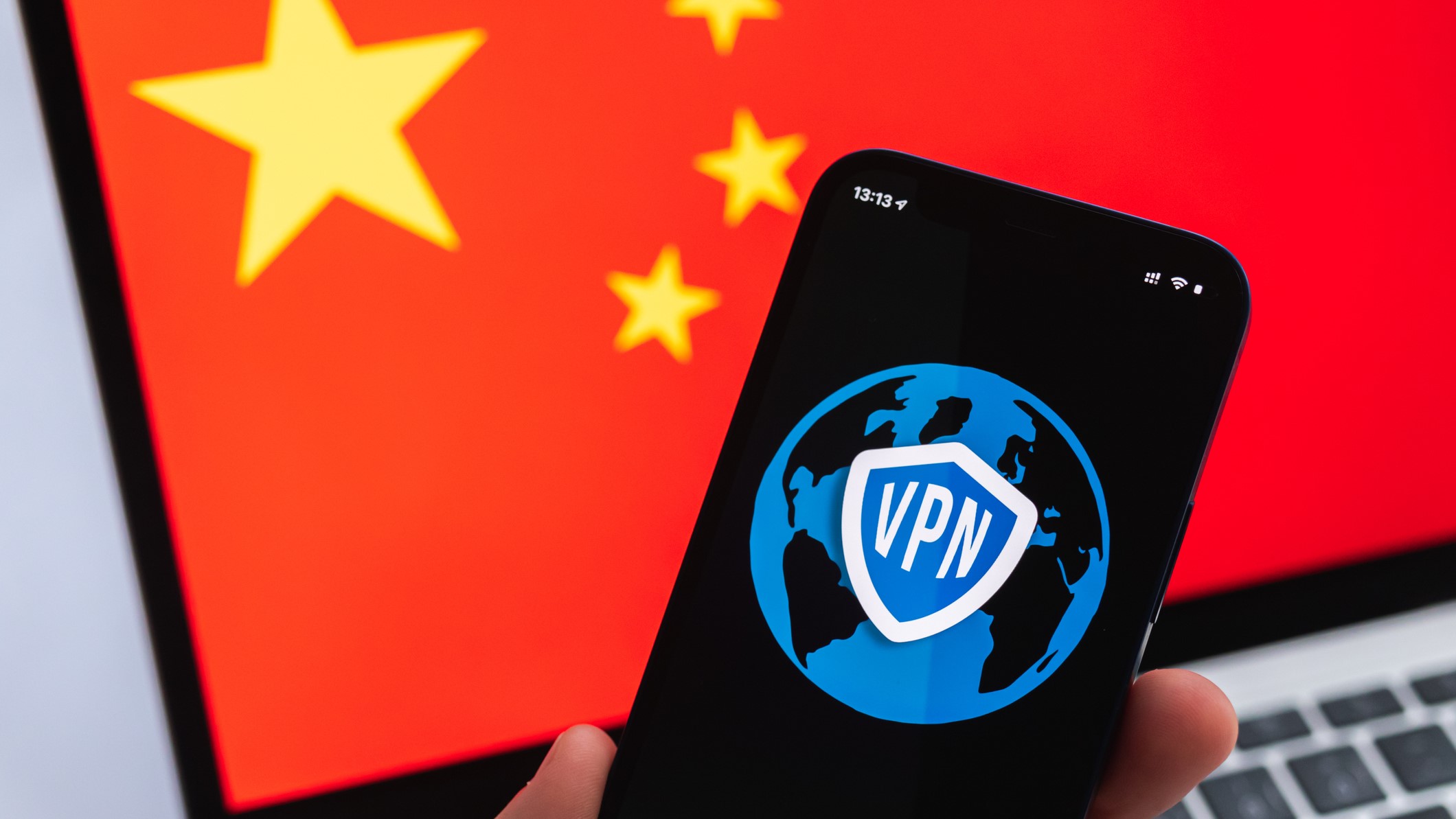 Hand with mobile phone and VPN app, Chinese flag on laptop screen in the background