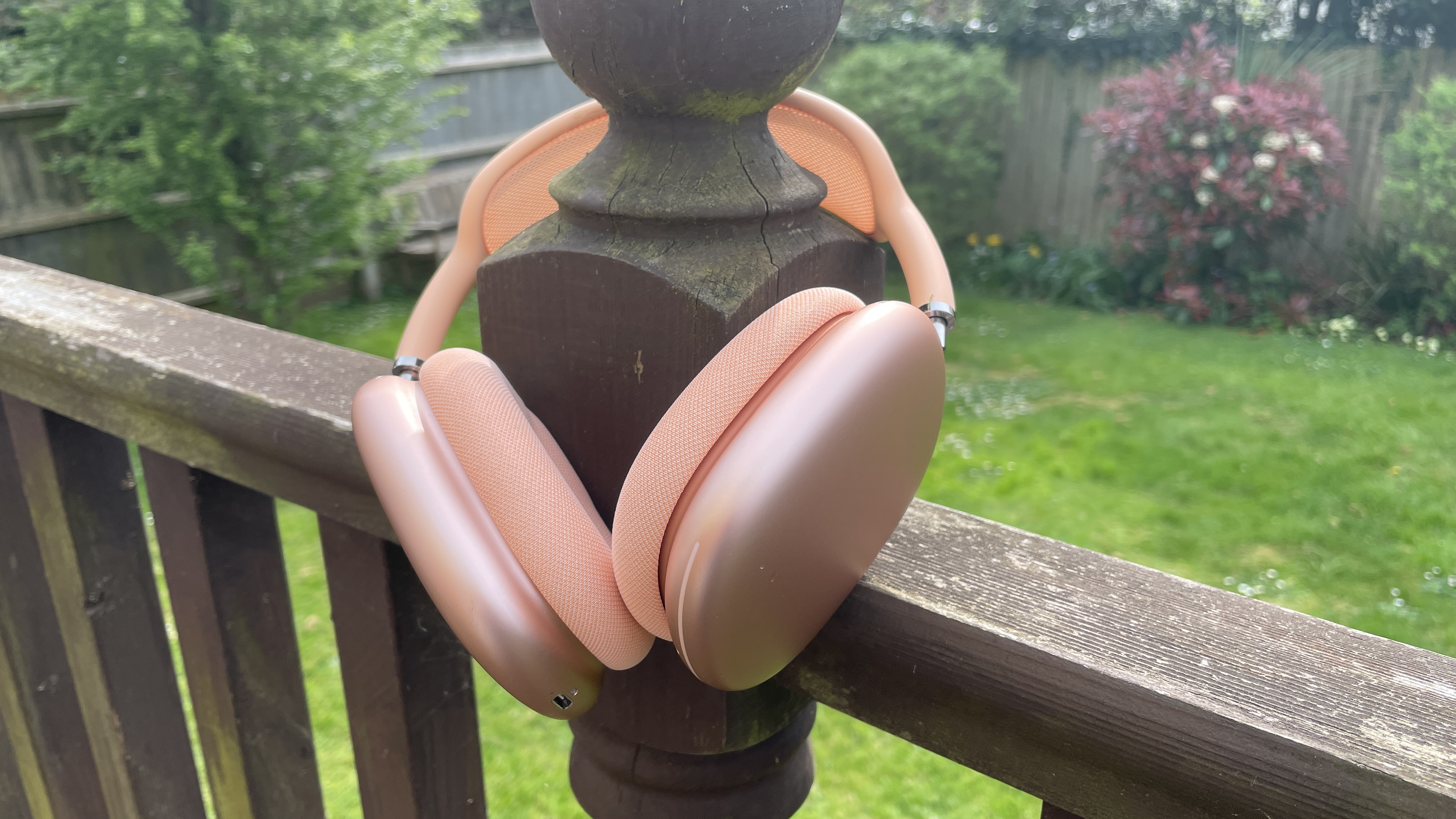 Apple AirPods Max 2 in orange with barely visible AirPods Max in purple, outside in a garden on a bench, held in one hand
