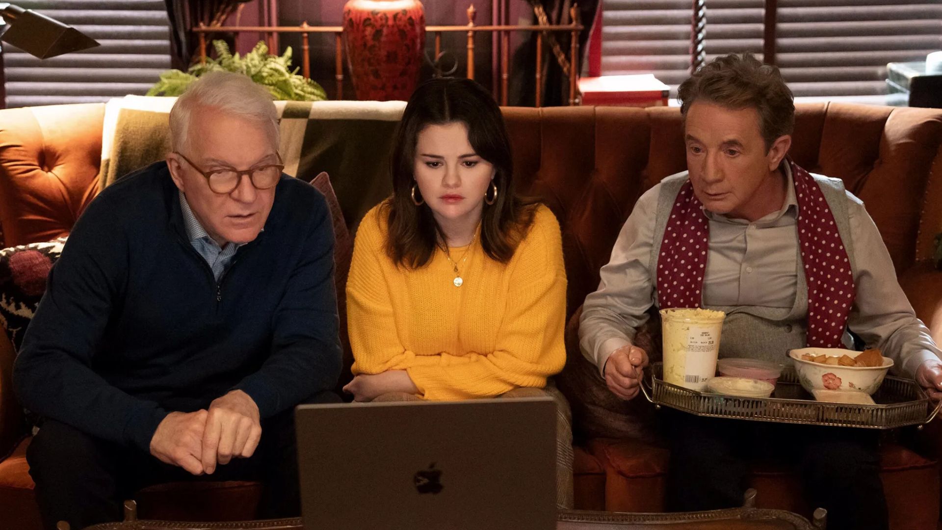 Steve Martin, Selena Gomez and Martin Short are sitting on a couch looking at a laptop, with a tray of food on Short's lap.