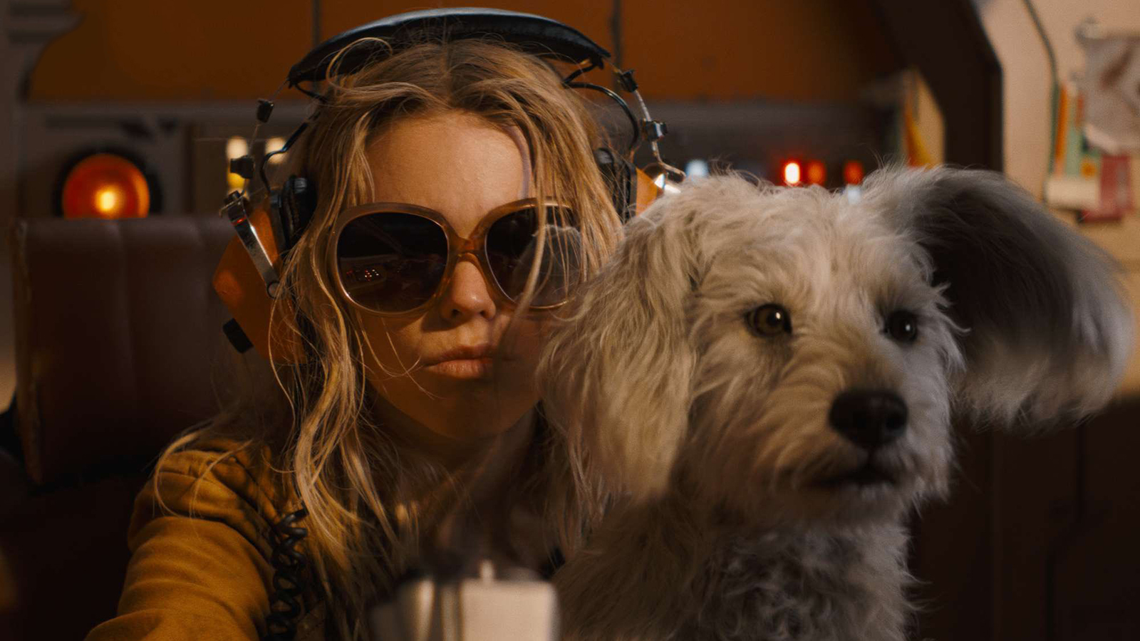 Kara wearing sunglasses and headphones while sitting in a booth with Krypto on her lap in Supergirl