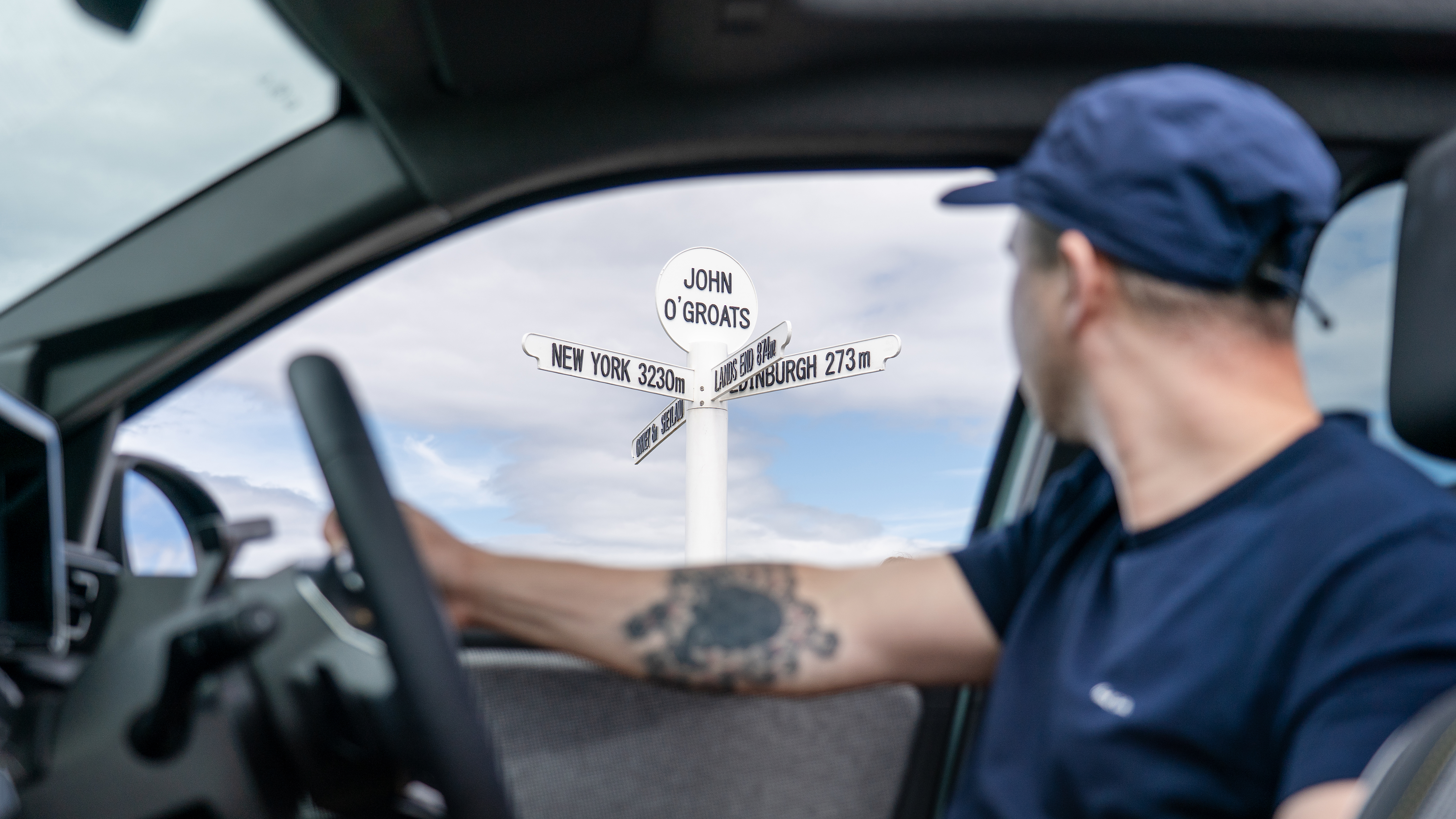 A man looking out the window of an electric vehicle at a sign indicating John O'Groats