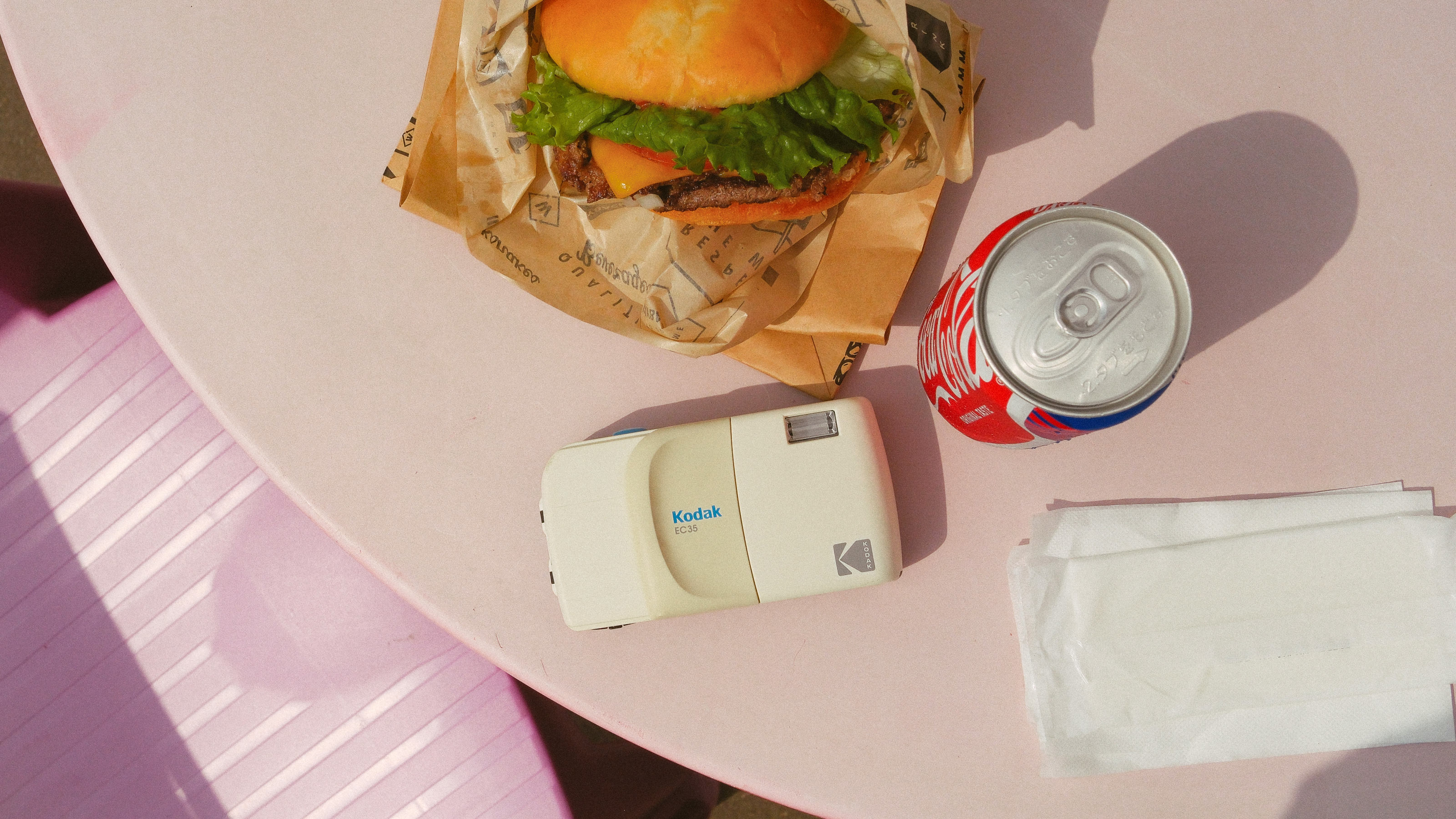 Photograph of blank Kodak EC35 film camera on a table next to a hamburger and a can of Coca-Cola