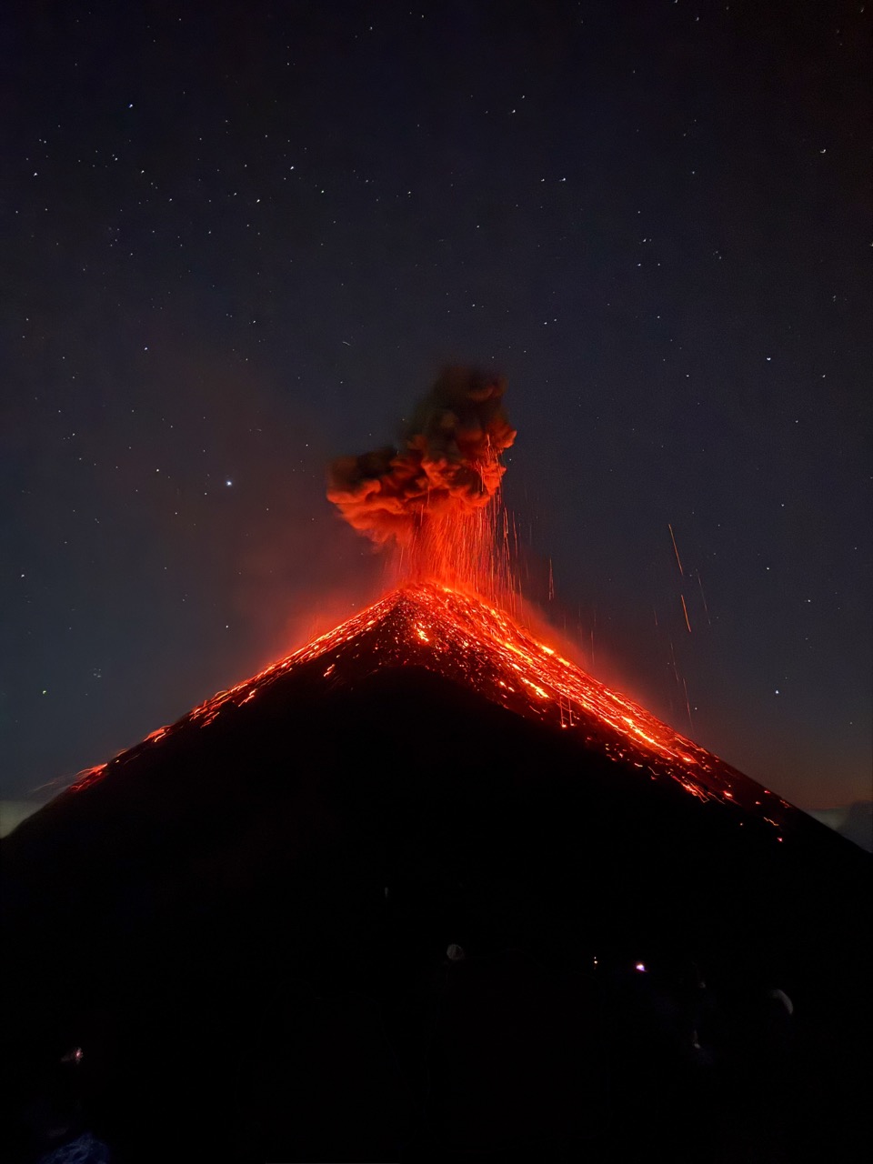 A photograph of a volcano fully erupting at night, with the bright red molten rock contrasting greatly with the mountainside shrouded in darkness and the night sky dimly lit by stars.