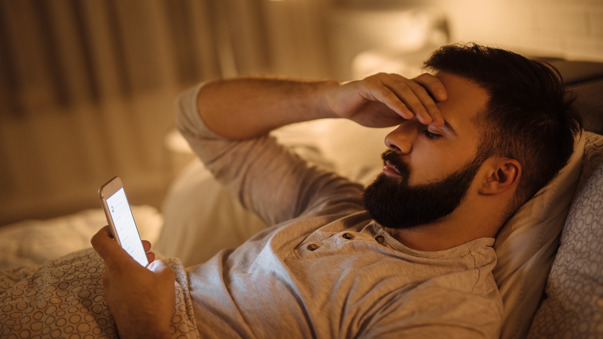 A young man is lying in bed looking at his phone, he looks stressed
